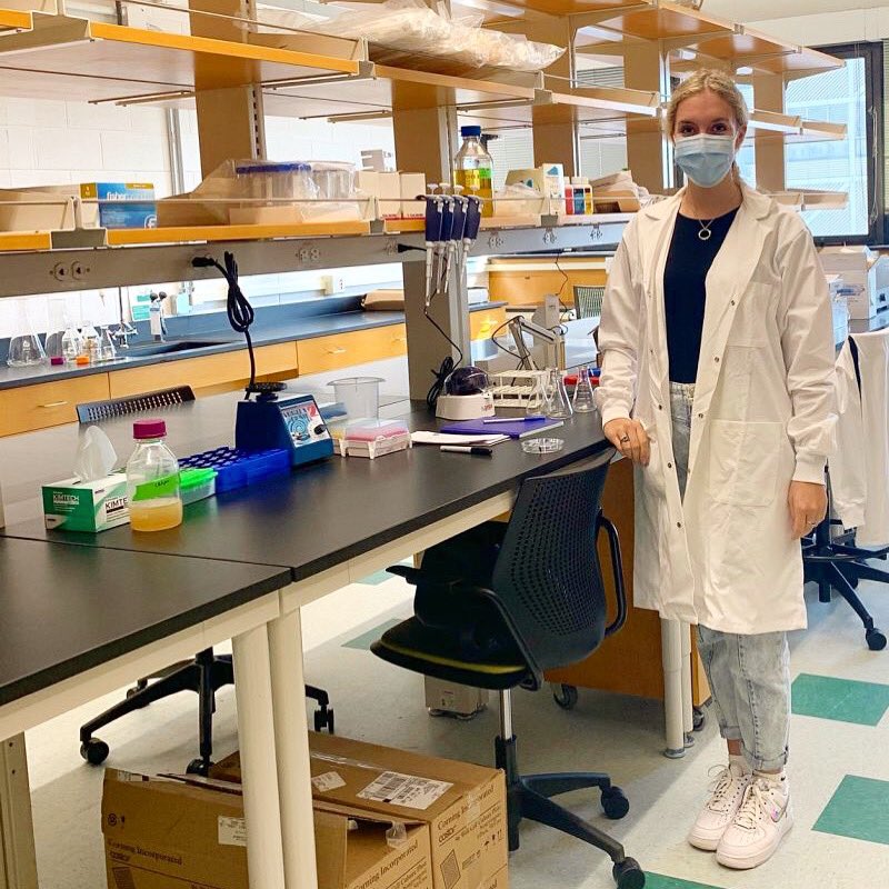 Student Hannah Prensky in a lab on campus, standing up wearing a white lab coat and a mask.